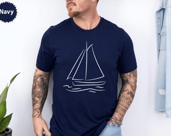 Peut inclure: T-shirt bleu marine avec un dessin au trait blanc d'un voilier. Le motif représente la coque, les voiles et les vagues. Le mot "Navy" est dans un cercle bleu marine en haut à gauche.