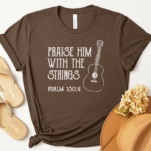 May include: Heather brown t-shirt with the text "Praise Him With The Strings" and a guitar illustration. Psalm 150:4 is printed below the text. The shirt is paired with beige sandals and a tan hat.