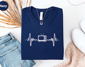 May include: Navy blue t-shirt with a white graphic of a vintage television set within a heartbeat line. A silver necklace with a sun pendant is visible. The shirt is folded on a white surface with blue jeans and white shoes.