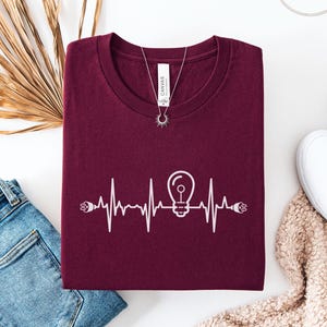 May include: A maroon t-shirt featuring a white graphic of a lightbulb within a heartbeat line. The shirt is styled with blue jeans, white trainers, and a necklace. The word "Maroon" is in a circular label in the upper left corner.