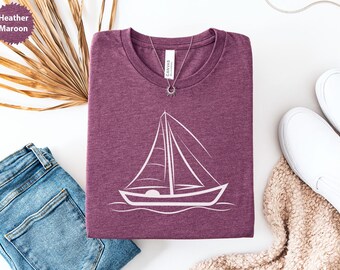 Peut inclure: T-shirt de couleur bordeaux avec un motif de voilier blanc. L'image comprend un jean bleu, des baskets blanches et un collier argenté. L'étiquette du t-shirt indique "Canvas".