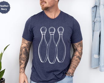 May include: Heather navy t-shirt with a white graphic of three bowling pins. The shirt has a crew neck and short sleeves. The text "Heather Navy" is in a navy blue circle in the upper left corner.