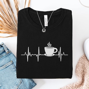 May include: Black t-shirt with a white graphic of a coffee cup and steam, integrated into a heartbeat line design. A silver necklace with a sun pendant is on the shirt. The shirt is on a white surface with blue jeans and white shoes.