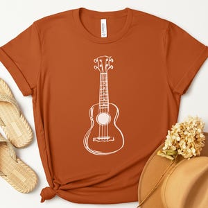 May include: Rust-colored t-shirt featuring a white ukulele outline. The shirt is paired with woven sandals, a tan hat, and dried flowers. The word "Autumn" is in a circular orange design in the upper left corner.