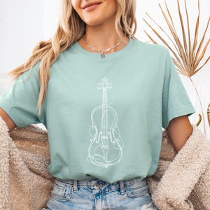 May include: A dusty blue t-shirt with a white line drawing of a violin. The shirt has short sleeves and a crew neck. The text "Dusty Blue" is in a light blue circle in the upper left corner.