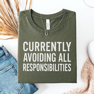 May include: Olive green t-shirt with the phrase "CURRENTLY AVOIDING ALL RESPONSIBILITIES" in white block letters. The shirt is laid flat with blue jeans, white sneakers, and a necklace.