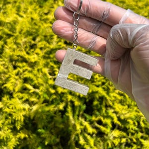 Op de afbeelding: Een betonnen letter "E" sleutelhanger met een zilveren ketting en sleutelring. De letter heeft een getextureerde, gemarmerde uitstraling. De achtergrond is een wazig beeld van groen gebladerte.