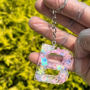 Op de afbeelding: Een heldere hars sleutelhanger in de vorm van de letter "B", gevuld met roze, witte en iriserende glitter, bloemen en schelpvormige versieringen. De sleutelhanger is bevestigd aan een zilveren ketting en sleutelring. De achtergrond is een wazig groen.