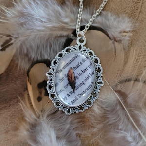 May include: Silver-toned pendant necklace with an oval-shaped charm. The charm features a printed text and a small feather. The pendant is set against a backdrop of brown and white feathers.