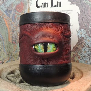May include: A cylindrical mug wrapped in textured dark red leather featuring a prominent, realistic green and yellow dragon eye at the center. The mug has smooth black leather bands at the top and bottom edges, creating a distinct contrast.