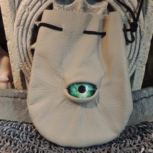 May include: A tan leather pouch with a large, green eye in the center. The pouch has a drawstring closure and is set against a backdrop of chainmail and a stone carving. A small statue is visible on the left.