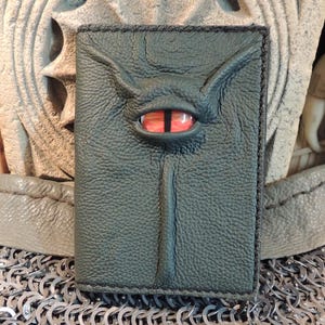May include: Dark green leather wallet with a red eye design. The wallet has a textured surface and a stitched border. The design includes a central vertical line and curved lines above the eye, giving it a unique, fantasy-inspired look.