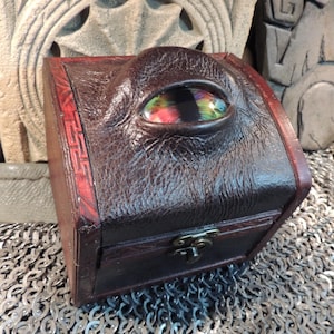 May include: A dark brown leather box with a decorative eye-shaped glass element. The box has a hinged lid and a metal clasp. The edges are trimmed with red leather. The box is sitting on a chainmail surface.