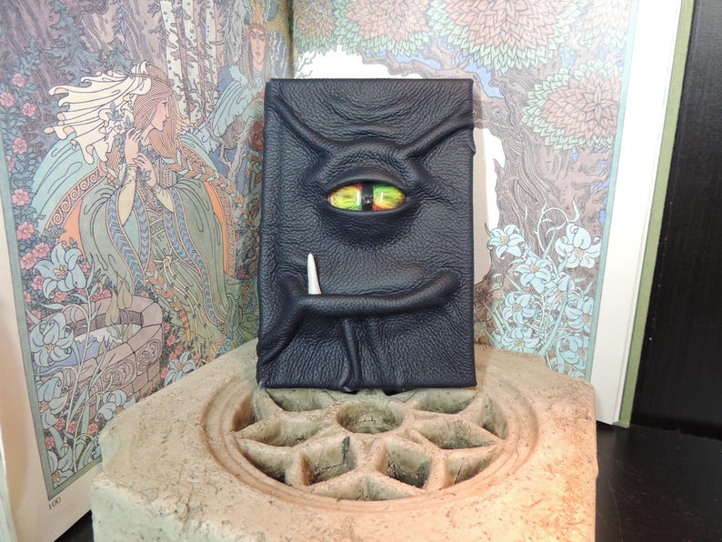 May include: A black leather journal with a textured surface, featuring a single, large, colorful eye and a white tusk. The journal is designed to resemble a monster face. The background includes illustrations from a book.