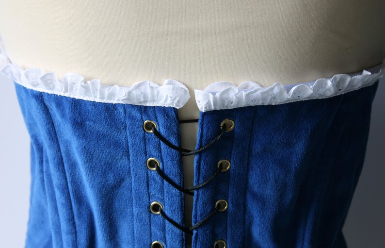 May include: Blue velvet corset with white lace trim and black lacing. The corset has gold metal eyelets and is tied at the back.