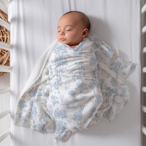 May include: A sleeping baby wrapped in a white and blue patterned swaddle blanket. The blanket features a repeating design of trees, giraffes, elephants, and other animals. The baby is lying in a white crib.