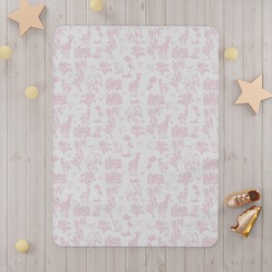 May include: A rectangular baby blanket with a pink safari animal pattern on a white background. The pattern includes giraffes, elephants, and trees. The blanket is surrounded by wooden stars, balls, and a pair of gold shoes.