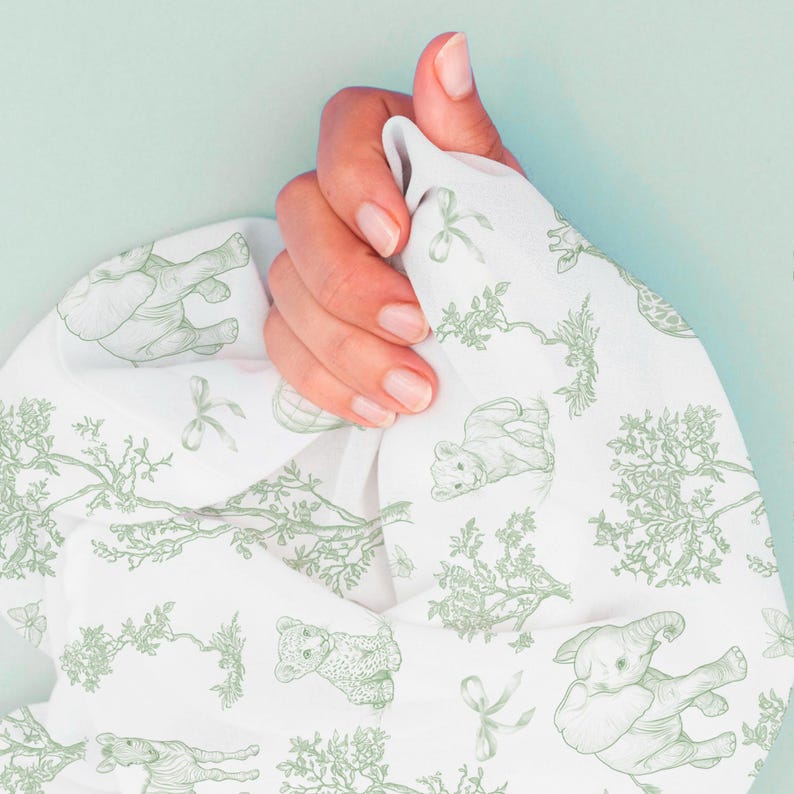 May include: White fabric with a repeating pattern of green elephants, bears, and trees. The fabric is held in a hand, with the design visible on the front. The background is a solid light blue.
