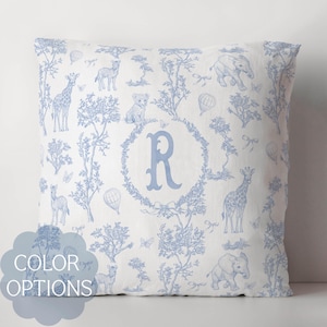Op de afbeelding: Vierkant sierkussen met een witte achtergrond en een blauw toile de Jouy-patroon met giraffen, olifanten en andere safaridieren. Een grote letter "R" is gecentreerd in een bloemenkrans. De woorden "COLOR OPTIONS" staan in een blauwe wolk.