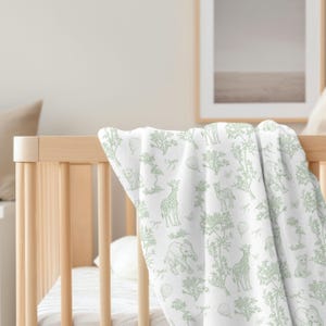 May include: A white baby blanket with a repeating pattern of green safari animals, trees, and hot air balloons. The blanket is draped over a wooden crib, showcasing the soft, detailed design.