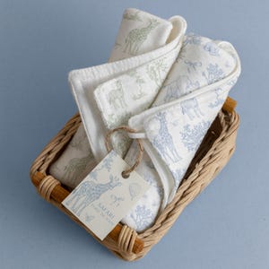 May include: A set of white baby washcloths with safari animal prints in a woven basket. The washcloths feature giraffe, elephant, and tree designs in green and blue. A tag with the words "Safari Toile de Jouy" is attached.