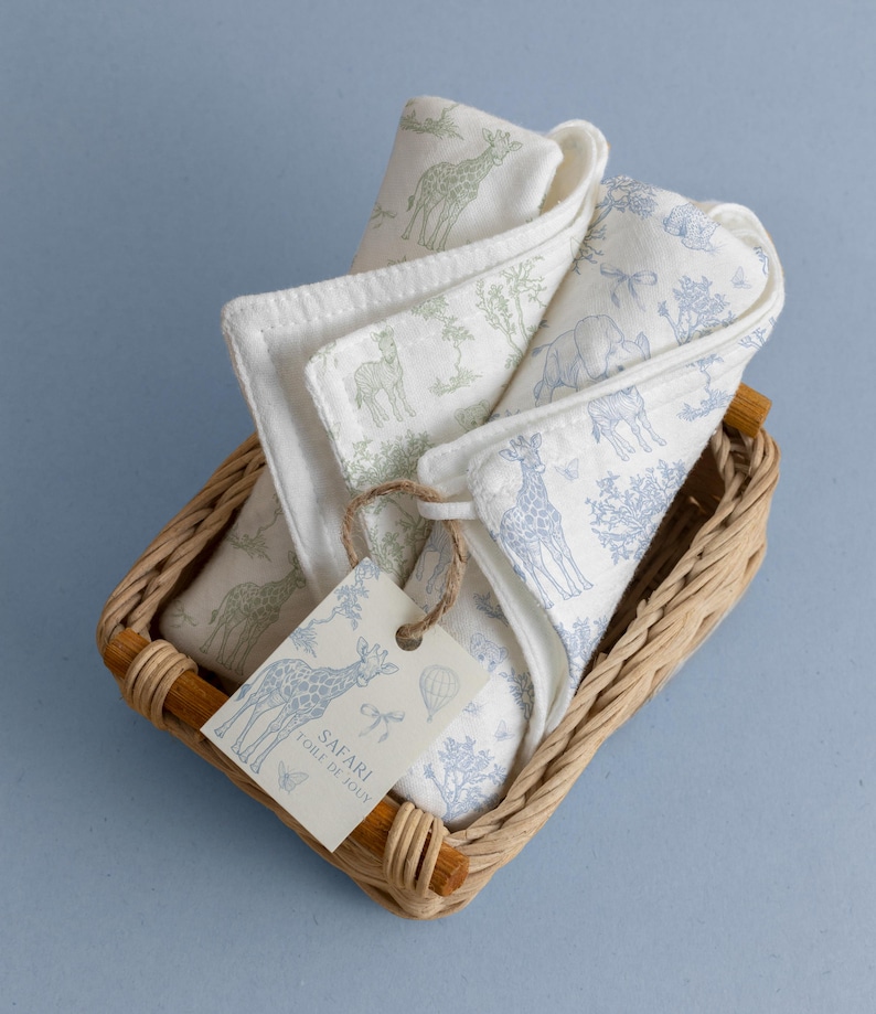 May include: A set of white baby washcloths with safari animal prints in a woven basket. The washcloths feature giraffe, elephant, and tree designs in green and blue. A tag with the words "Safari Toile de Jouy" is attached.