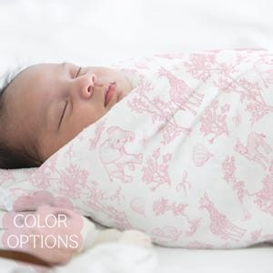 May include: A sleeping infant wrapped in a white and pink patterned swaddle blanket. The blanket features a repeating design of giraffes, elephants, and hot air balloons. The text "COLOR OPTIONS" is visible in the image.