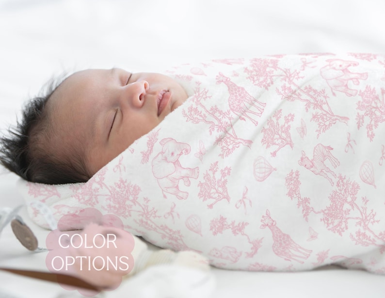 May include: A sleeping infant wrapped in a white and pink patterned swaddle blanket. The blanket features a repeating design of giraffes, elephants, and hot air balloons. The text "COLOR OPTIONS" is visible in the image.