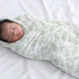 May include: A sleeping infant wrapped in a white and green patterned swaddle blanket. The blanket features a repeating design of giraffes, elephants, and trees. The baby has dark hair and is lying on a white sheet.