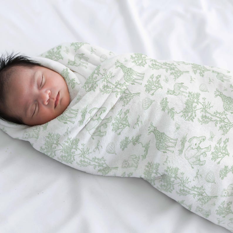 May include: A sleeping infant wrapped in a white and green patterned swaddle blanket. The blanket features a repeating design of giraffes, elephants, and trees. The baby has dark hair and is lying on a white sheet.