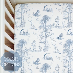 Puede incluir: Sábana bajera ajustable blanca con un estampado toile de Jouy azul con Winnie the Pooh, Piglet, Tigger y Eeyore. El diseño incluye árboles, globos e ilustraciones caprichosas. Las palabras "COLOR OPTIONS" están en una burbuja azul.