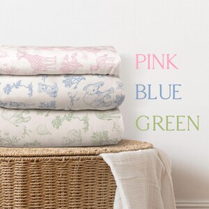 May include: A stack of three folded baby blankets with animal illustrations in pink, blue, and green. The blankets are on a woven basket with a white cloth draped over it. The words "PINK", "BLUE", and "GREEN" are written on the wall.