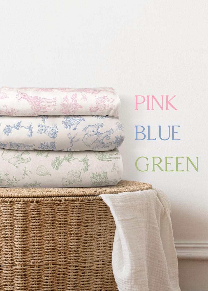 May include: A stack of three folded baby blankets with animal illustrations in pink, blue, and green. The blankets are on a woven basket with a white cloth draped over it. The words "PINK", "BLUE", and "GREEN" are written on the wall.
