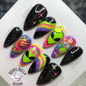 May include: A set of ten almond-shaped press-on nails featuring a space theme. Designs include black glitter backgrounds with colorful stars, neon green alien faces, rainbow swirls, and planetary illustrations. The logo Rebel Bella Dezigns is visible.