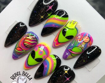 May include: A set of ten almond-shaped press-on nails featuring a space theme. Designs include black glitter backgrounds with colorful stars, neon green alien faces, rainbow swirls, and planetary illustrations. The logo Rebel Bella Dezigns is visible.