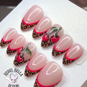 May include: A set of almond-shaped false nails. The nails feature a nude base with a pink French tip, a leopard print design, and cherry artwork. The logo "Rebel Bella Dezigns" is visible in the image.