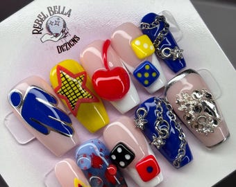 May include: A set of ten press-on nails featuring a nude base with various 3D embellishments including a red cherry, yellow and black dice, a yellow star, blue and silver chain charms, and clear rhinestones. The Rebel Bella Dezigns logo is printed.