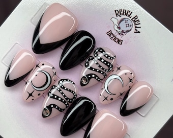 May include: A set of ten press-on nails featuring a glossy finish in black and nude. Designs include black French tips, a white crescent moon with stars, and a black snake with white dots. The Rebel Bella Dezigns logo is printed on the display card.