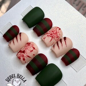 May include: A set of press-on nails with various designs. Some nails are solid dark green, while others feature red and green stripes or a blood splatter effect. The logo "Rebel Bella Dezigns" is visible.