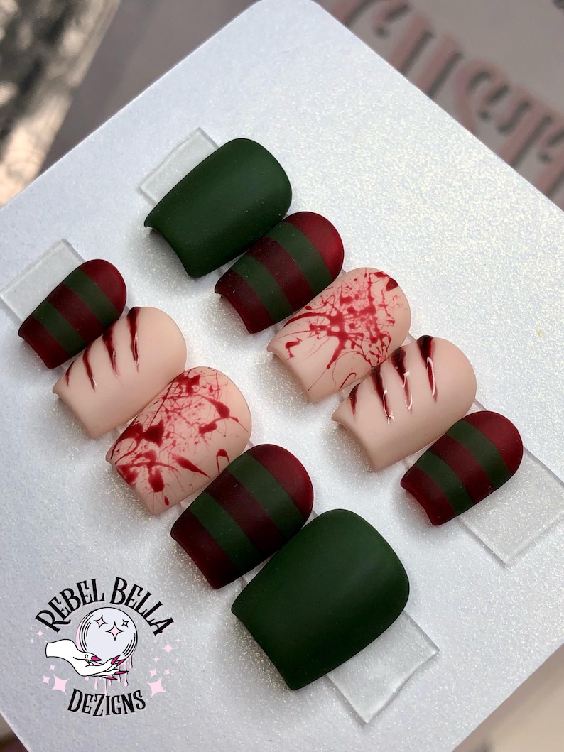 May include: A set of press-on nails with various designs. Some nails are solid dark green, while others feature red and green stripes or a blood splatter effect. The logo "Rebel Bella Dezigns" is visible.