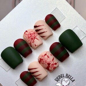 May include: A set of press-on nails with a horror theme. The nails feature designs including blood splatters, claw marks, and green and burgundy stripes. The logo "Rebel Bella Dezigns" is visible in the bottom right corner.