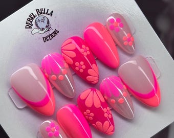 Könnte beinhalten: Ein Set von künstlichen Nägeln in verschiedenen Rosa- und Koralltönen. Einige Nägel weisen florale Designs auf, andere sind einfarbig oder haben eine French-Spitze. Das Markenlogo "Rebel Bella Dezigns" ist sichtbar.