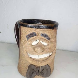 May include: A whimsical, handmade ceramic mug with a smiling face design. The mug features a brown base color with a dark brown rim, handle, and bow tie. The face has white teeth and eyes, and the mug is approximately 5 inches tall.