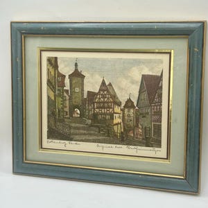 May include: Framed print of a European town scene with buildings and a tower. The print is in a teal and gold frame. The print has a light green matting.