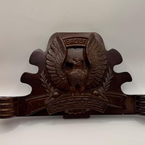 May include: A dark brown wooden wall-mounted pipe rack featuring a carved eagle with outstretched wings. The word "EAGLE" is carved above the eagle. The rack has two cylindrical holders and a decorative banner below the eagle.