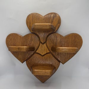 May include: A wall-mounted wooden display shelf featuring four interconnected heart-shaped sections, each with a small horizontal ledge. The wood has a medium-brown stained finish with a visible grain pattern, arranged in a clover-like configuration.