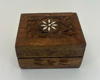 May include: A small, square wooden box with a hinged lid. The box is a warm brown color and features carved floral designs. The lid has a central white flower inlay. The box is likely used for storing small items like jewelry.