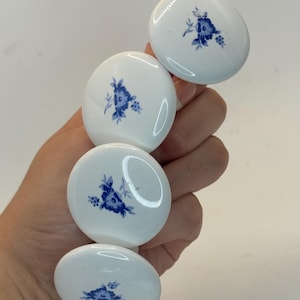 May include: Four white ceramic cabinet knobs with a blue floral design. The knobs are oval-shaped and held in a hand. The floral pattern is a delicate blue, adding a touch of elegance to the knobs.