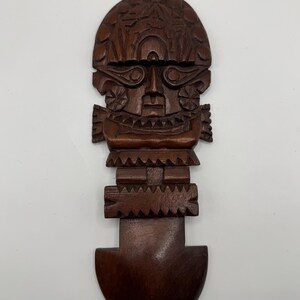 May include: A dark brown, carved wooden tiki figure with a stylized face, intricate details, and a rounded base. The figure has a headdress and a rectangular body with geometric patterns. The wood has a natural grain.