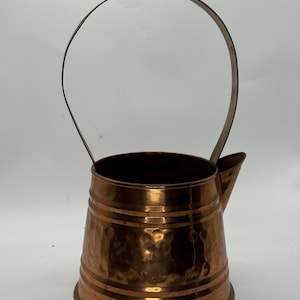 May include: A copper-colored pitcher with a handle and a spout. The pitcher has a hammered texture and horizontal bands around the body. The handle is made of a darker metal and arches over the top.